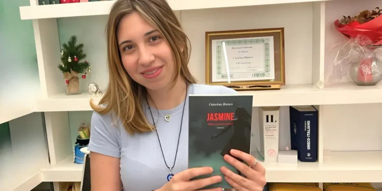 Caterina Bianco sorride tenendo in mano il suo romanzo d’esordio “Jasmine, attiva l’aura emotiva!” davanti a una libreria.